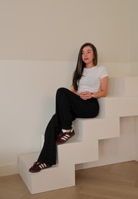 Hvit kortermet skjorte, svarte vide bukser og maroon sneakers med hvite striper. Sitter på hvite geometriske trinn. Nøytral bakgrunn.