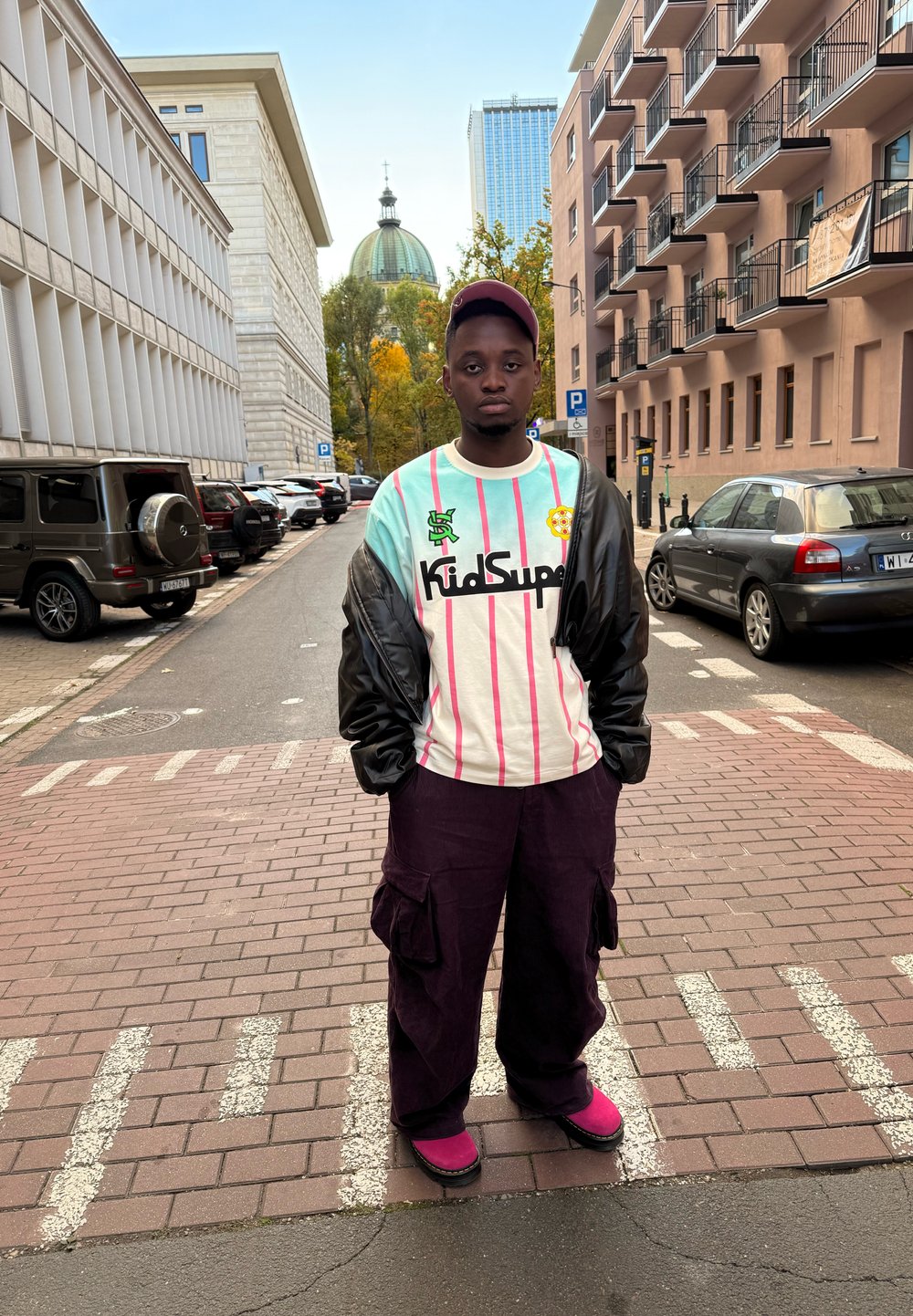 Person wearing an oversized light blue and white striped shirt with black detailing, purple cargo trousers, and pink footwear, standing on a street.
