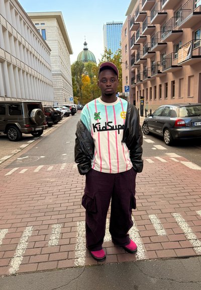 Persona vestida con una camisa de rayas azul claro y blanca de gran tamaño con detalles en negro, pantalones cargo morados y calzado rosa, de pie en una calle.