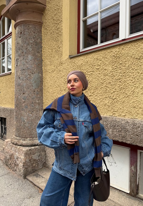 Mujer con pañuelo marrón en la cabeza, chaqueta vaquera azul y bufanda a rayas, de pie junto a una pared amarilla texturizada con ventanas y una columna de piedra.