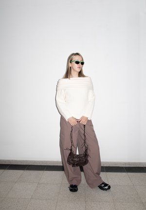 Mujer con suéter blanco de hombros descubiertos, pantalones marrones holgados, zapatos negros y gafas de sol verdes, sosteniendo un bolso pequeño negro contra una pared blanca.