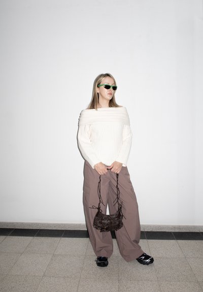 Mujer con suéter blanco de hombros descubiertos, pantalones marrones holgados, zapatos negros y gafas de sol verdes, sosteniendo un bolso pequeño negro contra una pared blanca.