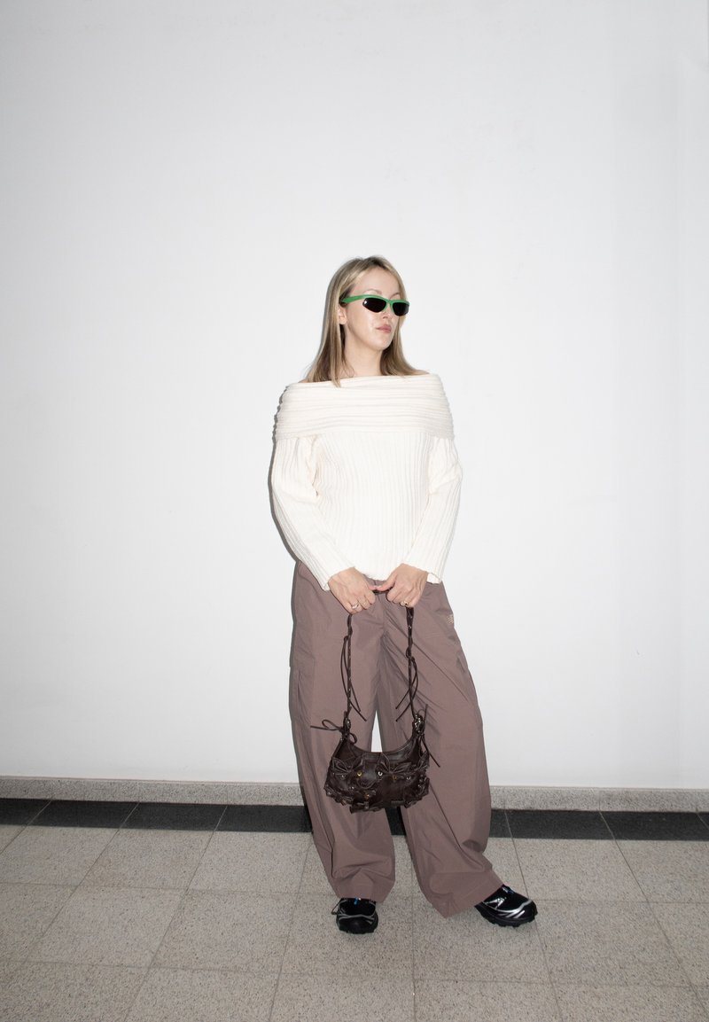 Femme portant un pull blanc à épaules dénudées, un pantalon marron ample, des chaussures noires et des lunettes de soleil vertes, tenant un petit sac à main noir contre un mur blanc.