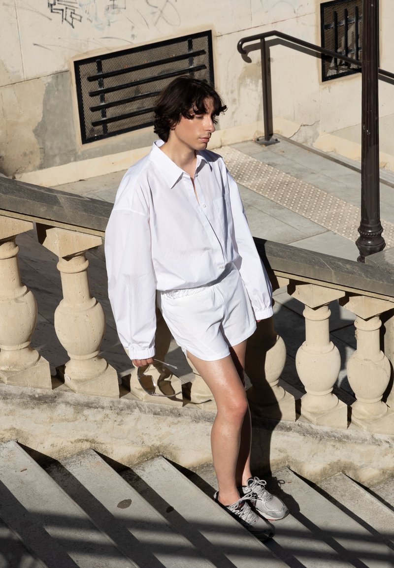 Personne aux cheveux courts et foncés portant une chemise blanche oversize, un short blanc et des baskets descendant des escaliers en pierre à l'extérieur sous un soleil éclatant.