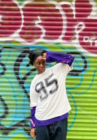 Camisa branca oversized com o número "95" em preto, sobreposta a uma camisa de mangas compridas roxa com detalhes em rosa. Jeans escuros, posado contra um fundo de graffiti colorido.