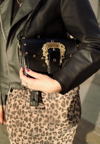 Bolso de cuero negro con herrajes de león dorados y embellecimientos de estrellas, sostenido por una persona que lleva una chaqueta de cuero negra y una falda de estampado de leopardo.
