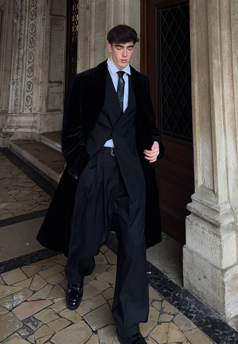 Manteau en velours noir, gilet croisé, chemise blanche habillée, cravate noire, pantalons noirs amples et chaussures noires brillantes.