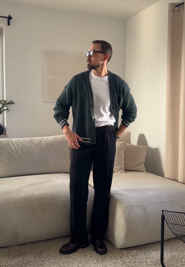 Homme portant des lunettes, un cardigan vert foncé, un t-shirt blanc et un pantalon noir, debout dans un salon beige près d'un canapé.