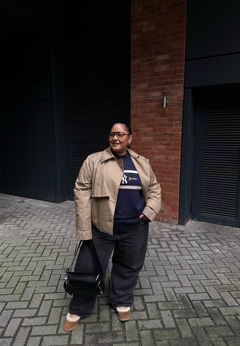 Personne portant une veste beige et un pantalon sombre, tenant un sac à main noir, debout sur une rue pavée à côté de murs en briques et d'un bâtiment sombre, souriant et regardant sur le côté.