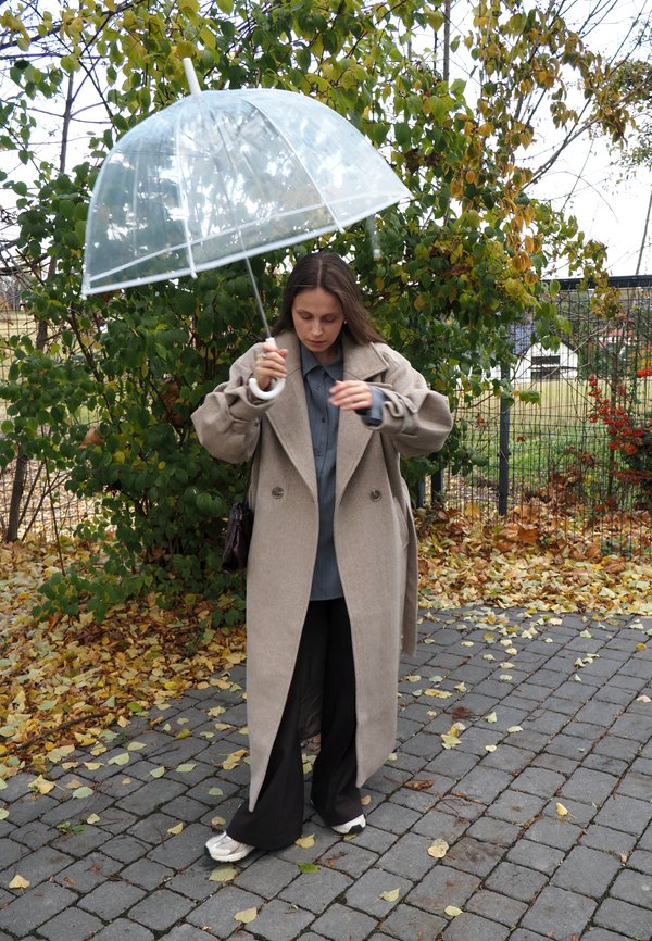 Beige Trenchcoat, graues Hemd mit Knöpfen, schwarze weite Hose und durchsichtiger Regenschirm. Umgeben von grünem Laub und gefallenen Blättern.