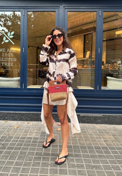 Camisa de manga larga con patrón de tie-dye en negro y beige, combinada con una falda beige, chanclas negras y un bolso de mano de mimbre con detalles en rojo y beige.