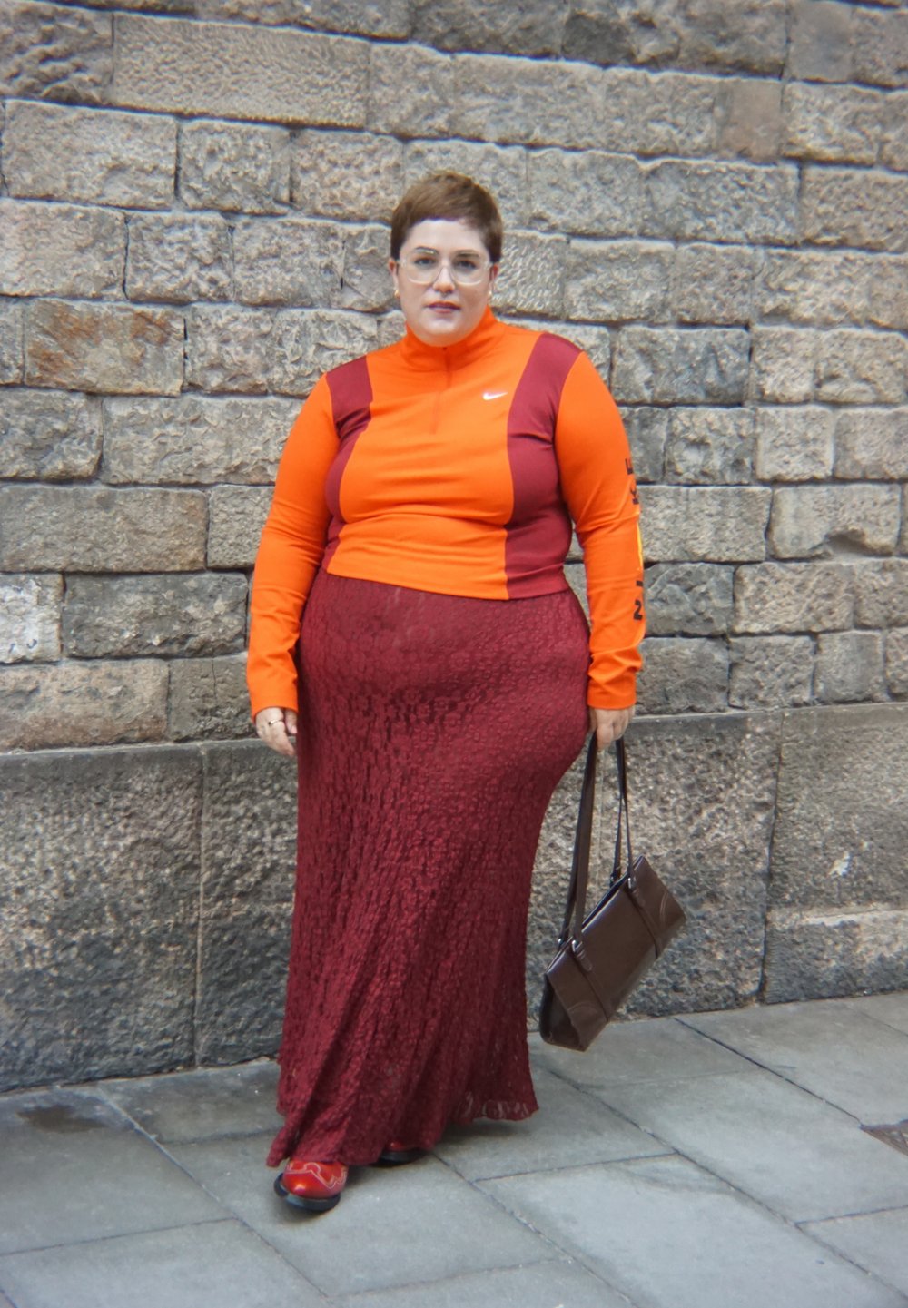 Dlhý top s dlhým rukávom s oranžovými a bordovými farebnými blokmi a stojatým golierom, v kombinácii s prúdiacou bordovou maxi sukňou s textúrou a hnedou kabelkou.