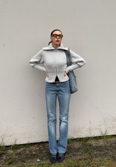Mujer con suéter de punto gris con cremallera, jeans azules, zapatos negros, gafas con lentes amarillas y llevando un bolso azul, de pie contra una pared lisa.