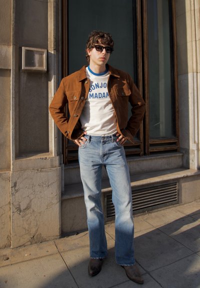 Chaqueta de ante marrón sobre una camiseta blanca con texto azul, jeans acampanados de color azul claro y botas marrones oscuras, contra un fondo de pared de piedra.