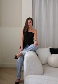Črna brezrokavna majica z gumbi spredaj, svetlo modri jeans in črni špicasti petki. Sedi na teksturirani svetle barve sedežni garnituri z blazinami.