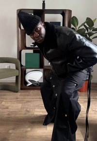 Veste en cuir noire avec col en fourrure, associée à un pantalon noir ample avec des pressions. La personne porte un bonnet tricoté et est à l'intérieur.