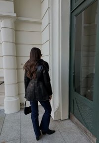 Črna usnjena jakna z ovratnikom iz krzna, temno modre kavbojke in črni gležnarji; stoji ob zeleni vratih in belih stebrih.