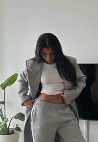 Kvinna med långt mörkt hår justerar grå sweatpants och vit skjorta under en grå kappa i ett minimalistiskt rum med en växt och TV.