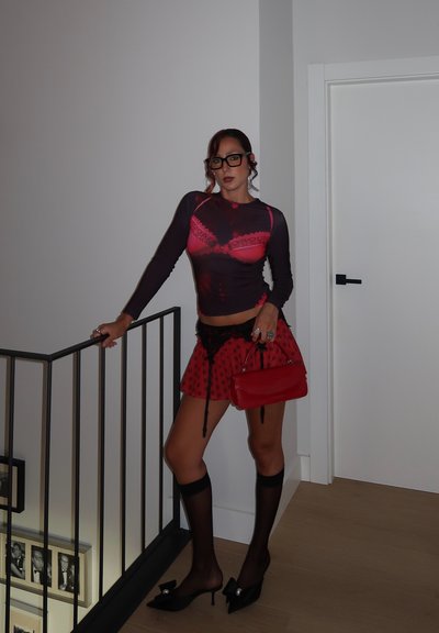 Mujer que lleva una blusa de manga larga transparente con estampado de sostén rojo, falda negra de lunares rojos, medias altas negras transparentes hasta la rodilla, tacones negros y sostiene un bolso rojo.