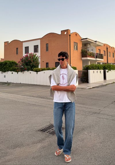 Suéter gris colgado sobre una camiseta blanca, jeans azules y chanclas blancas. Las gafas de sol complementan el look informal frente a un fondo de edificio de ladrillo.