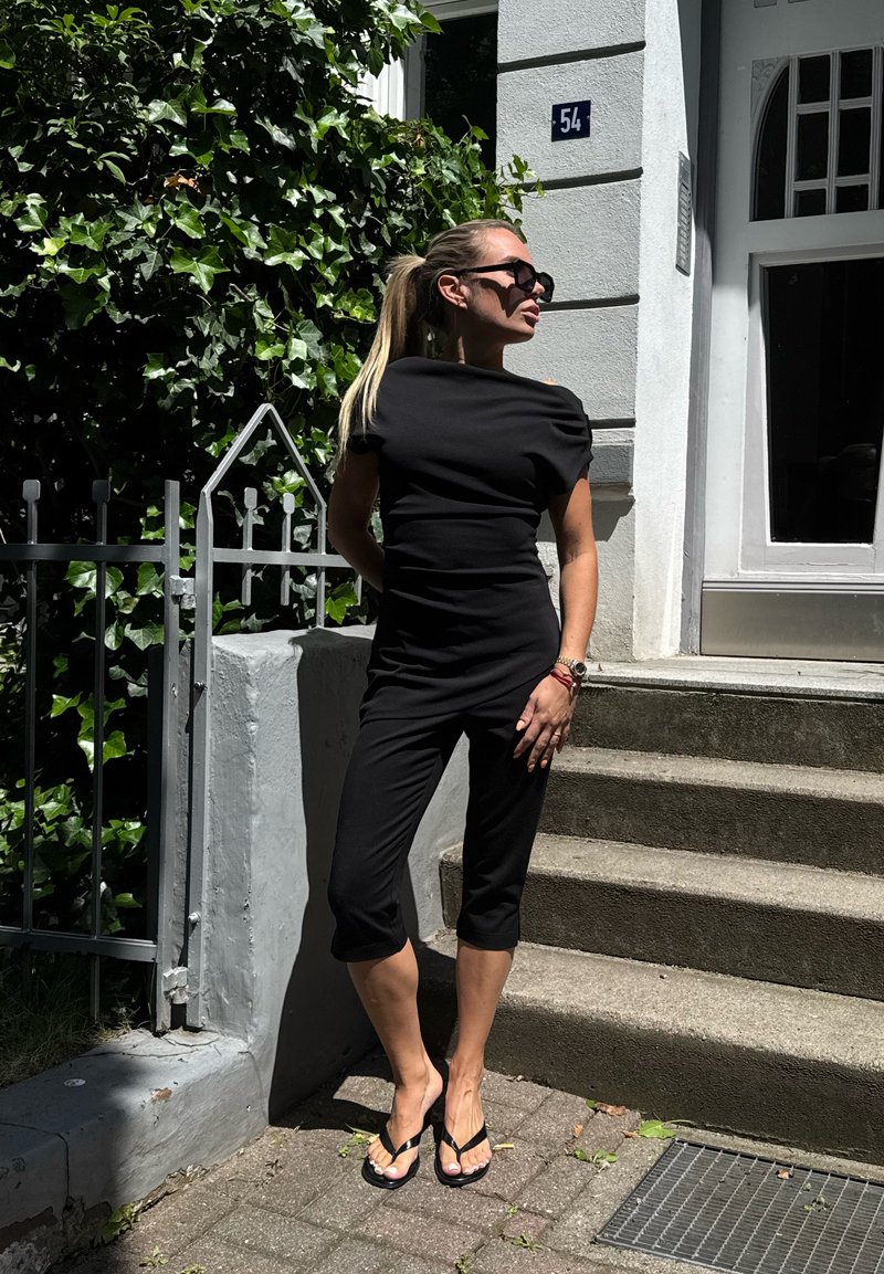 Femme portant une combinaison noire et des lunettes de soleil debout près d'escaliers et d'une porte avec du lierre vert par une journée ensoleillée.