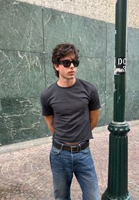 Ung mand med mørkt hår og solbriller, iført en sort t-shirt og blå jeans, stående med hænderne bag ryggen nær en grøn lygtepæl og en marmormur.