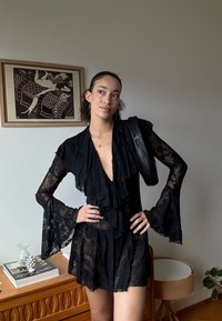 Robe en dentelle noire avec des détails volantés, des manches longues évasées et un col en V profond. En arrière-plan, une œuvre d'art et un meuble.