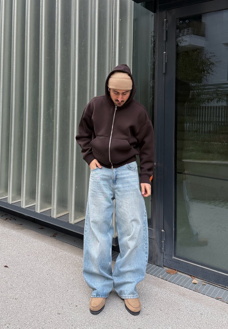 Homme portant un sweat à capuche marron, un bonnet beige, un jean large bleu clair, et des chaussures marron clair, debout à l'extérieur près d'une façade de bâtiment en verre et métal.