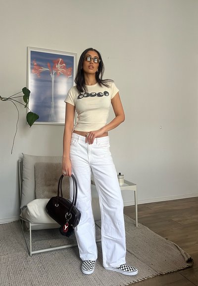Camiseta corta crema con diseño gráfico negro, jeans anchos blancos, bolso negro, zapatillas de cuadros, gafas de sol y pose informal.