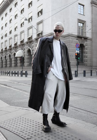 Persona con cabello corto y blanco que lleva un abrigo de cuero negro de gran tamaño, camisa blanca, pantalones gris claro, botas negras y gafas de sol en una esquina de la calle de la ciudad.