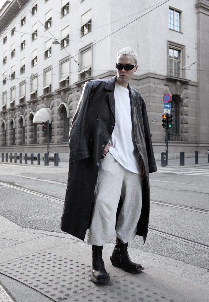 Personne aux cheveux blancs courts portant un manteau en cuir noir oversize, une chemise blanche, un pantalon gris clair, des bottes noires et des lunettes de soleil à un coin de rue en ville.