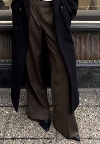 Pantalon marron à jambes larges en tissu lisse, associé à un manteau noir. La tenue est complétée par des chaussures noires à bouts pointus pour un look élégant.