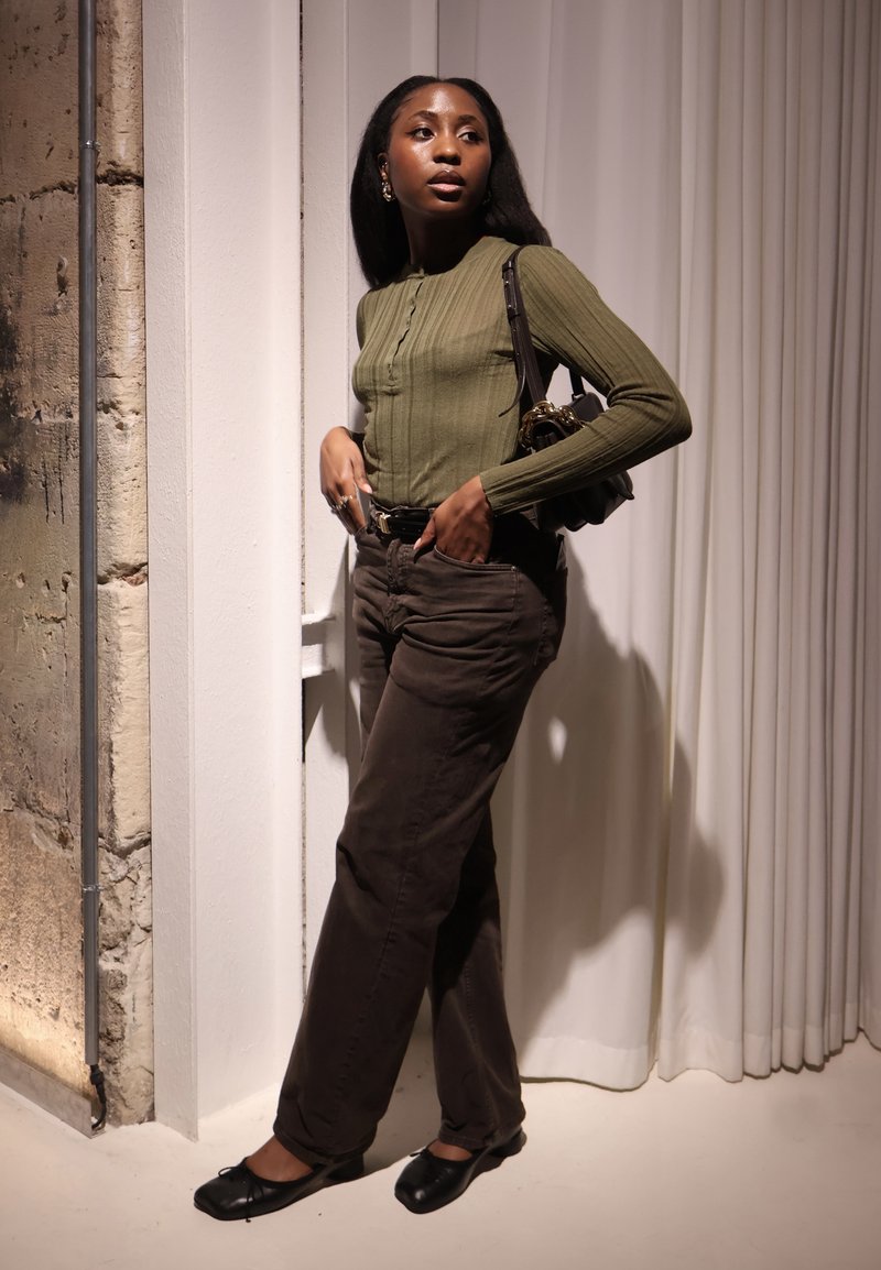 Haut vert côtelé à manches longues, pantalon marron foncé et ballerines noires. Le mannequin se tient près d'un mur texturé, tenant un petit sac, avec les cheveux raides.