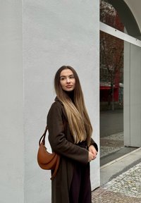 Jeune femme aux longs cheveux bruns portant un manteau foncé et un sac à bandoulière marron se tient près d’un mur blanc en extérieur.