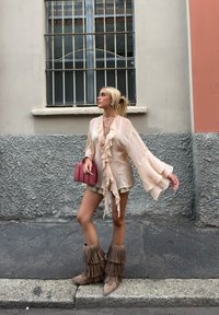 Blusa de gasa beige con volantes, combinada con pantalones cortos dorados. Lleva botas beige con flecos y sostiene un pequeño bolso rosa. Fondo de pared texturizada en gris.