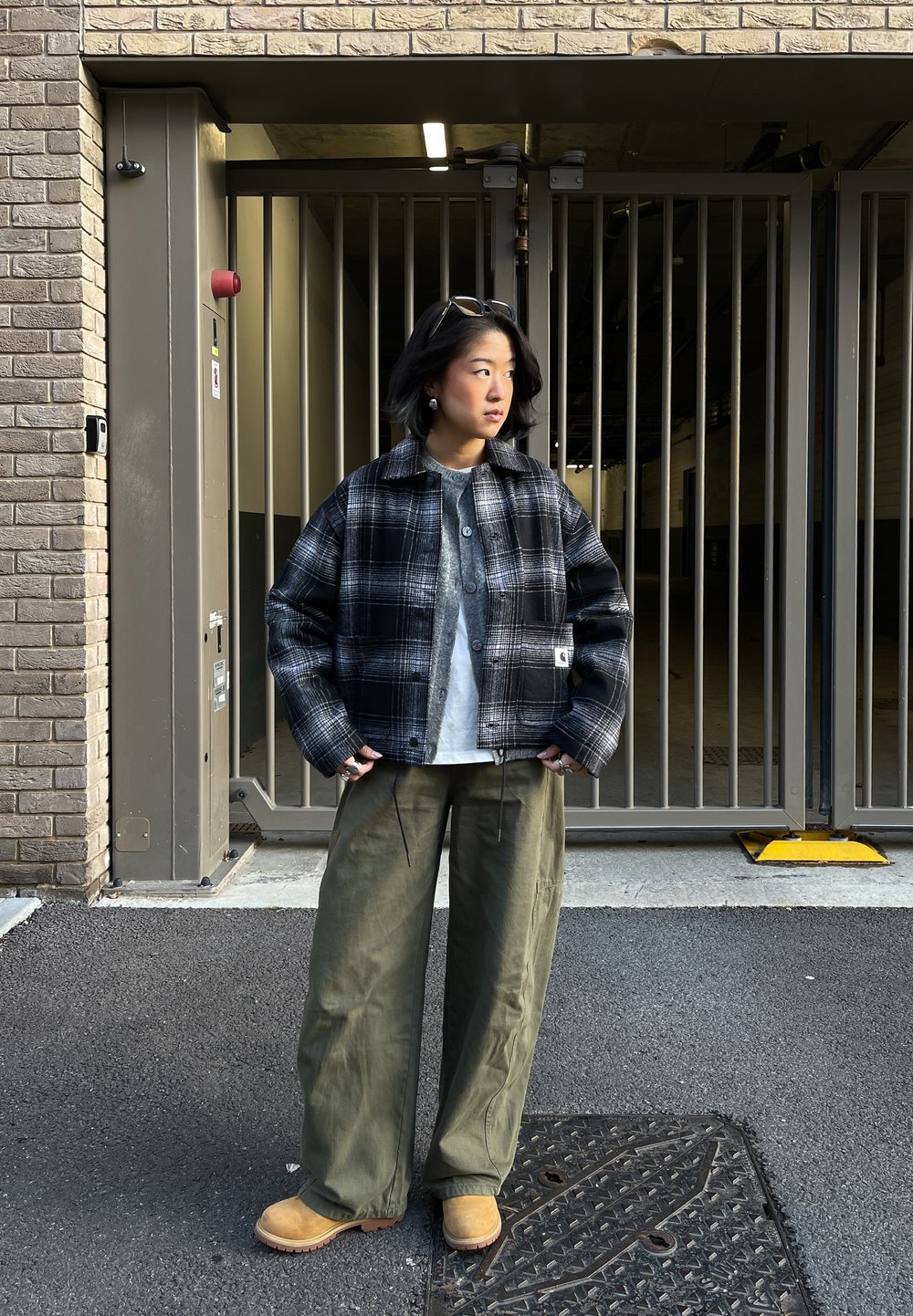 Personne portant une veste à carreaux noir et blanc, une chemise grise, un pantalon large vert et des bottes couleur sable, se tenant devant une grille en métal et un mur en briques.