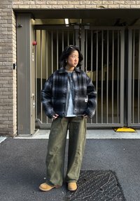 Personne portant une veste à carreaux noir et blanc, une chemise grise, un pantalon large vert et des bottes couleur sable, se tenant devant une grille en métal et un mur en briques.