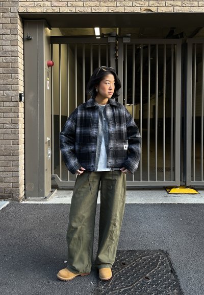 Persona con chaqueta de cuadros en blanco y negro, camisa gris, pantalones anchos verdes y botas color canela, de pie frente a una puerta metálica y una pared de ladrillos.
