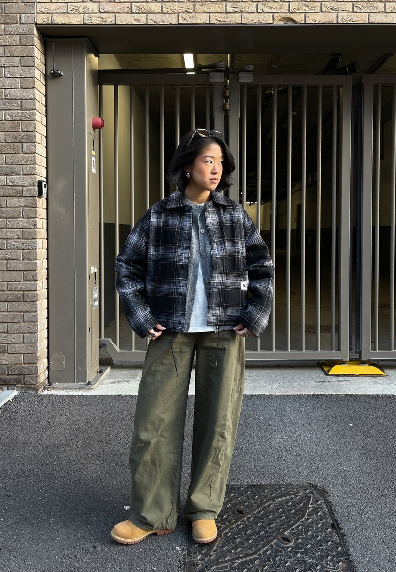 Personne portant une veste à carreaux noir et blanc, une chemise grise, un pantalon large vert et des bottes marron clair se tient devant une grille en métal et un mur de briques.
