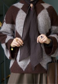 Pulover voluminos și confortabil în nuanțe de maro și gri, cu model în romburi; eșarfă închisă la culoare legată la gât; mai multe inele și brățară aurie pe mâini.