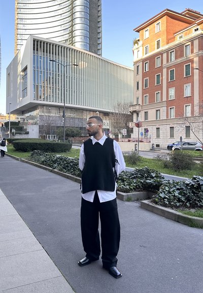 Chaleco de punto negro superpuesto a una camisa blanca con cuello, combinado con pantalones negros de pierna ancha y zapatos negros, con un fondo de arquitectura urbana.