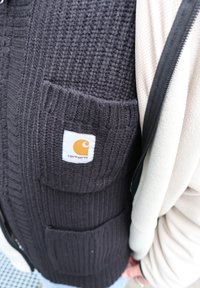 Gros plan sur une personne portant un gilet en maille noire de Carhartt par-dessus une veste beige clair zippée avec des poches visibles et un tissu texturé.