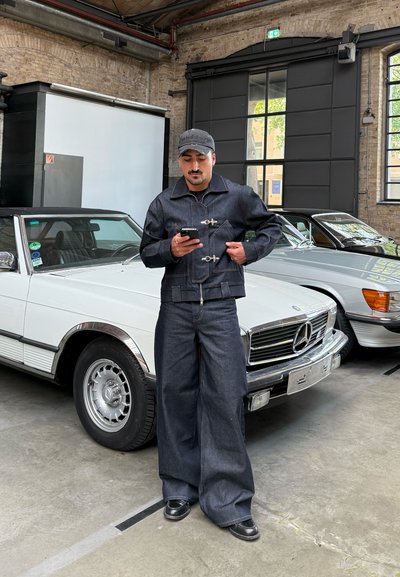 Hombre vestido con un conjunto de denim oscuro con cierre de cremallera y botón, de pie junto a un coche convertible blanco clásico en un entorno industrial.