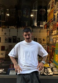 Tricou alb supradimensionat cu mâneci scurte, purtat peste pantaloni scurți negri. Tatuaje pe antebrațe vizibile. Vitrina magazinului afișează mărfuri și postere cu formații.