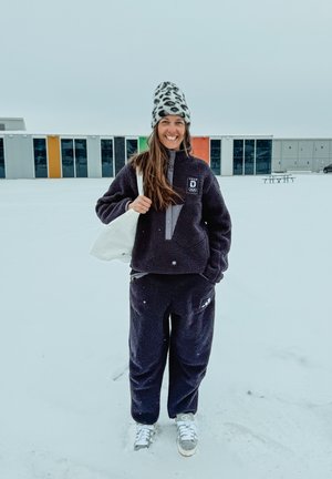 Mujer sonriente con conjunto de forro polar negro y gorro de leopardo de pie en la nieve con una bolsa blanca sobre el hombro, edificio colorido al fondo.