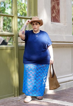 Camiseta de manga corta azul marino, falda larga de encaje azul, sombrero beige, bolso marrón, sandalias, tatuajes visibles en el brazo. Marco de ventana verde en el fondo.