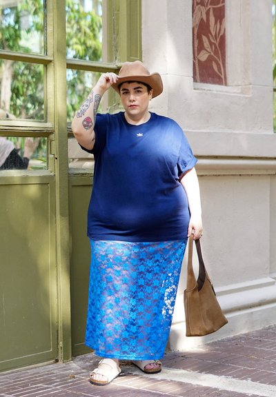 Camiseta de manga corta azul marino, falda larga de encaje azul, sombrero beige, bolso marrón, sandalias, tatuajes visibles en el brazo. Marco de ventana verde en el fondo.