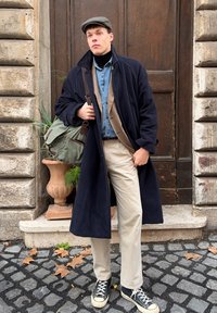 Un jeune homme portant un manteau bleu marine, un pantalon beige, des baskets noires et une casquette plate se tient contre une porte en bois sur une rue pavée, avec un sac vert.