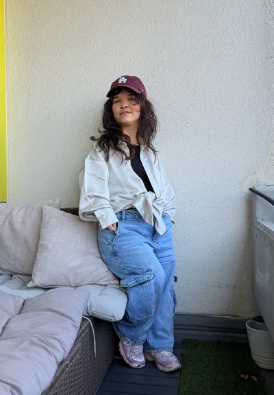 Joven con cabello oscuro que lleva una gorra burdeos, camisa blanca, camiseta negra, vaqueros cargo azules y zapatillas, apoyada contra una pared beige en un balcón.