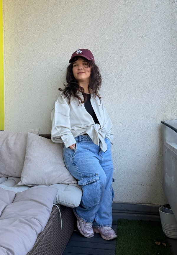 Giovane donna con capelli scuri che indossa un berretto color vinaccia, una camicia bianca, una maglietta nera, jeans cargo blu e sneakers, appoggiata a un muro beige su un balcone.
